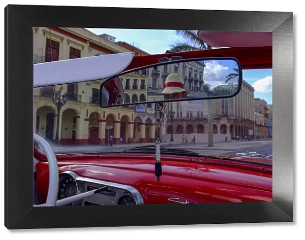 Taxi driver in straw hat seen in rear-view mirror of vintage car, Havana, Cuba