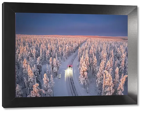 Aerial view of car on icy road and illuminated headlamps driving in the snowcapped forest, at dawn, Akaslompolo, Kolari, Pallas-Yllastunturi National Park, Lapland region, Finland, Europe