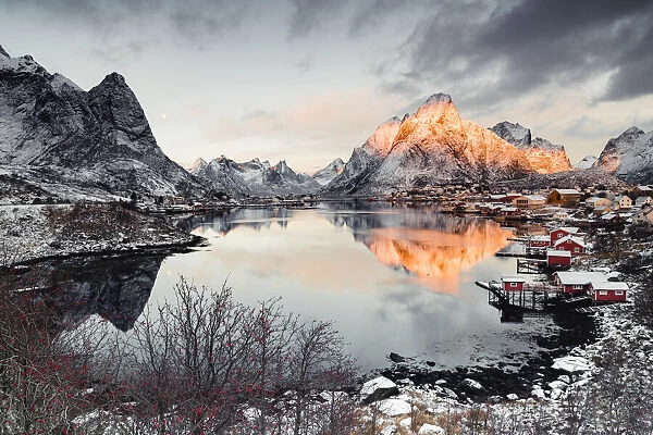 Clouds at dawn over traditional Rorbu and Olstind mountain reflected in sea, Reine Bay