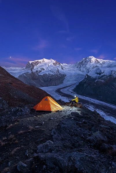Hiker with head lamp sitting beside the tent in front of Monte Rosa massif, Gorner glacier (Gornergletscher) and Lyskamm peaks at dusk, Riffelsee, Zermatt, canton of Valais, Swiss Alps, Switzerland, Europe