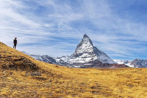 Hiker stands in front of the iconic shape of Matterhorn among yellow grasses, Riffelalp, Zermatt, Valais Canton, Swiss Alps, Switzerland, Europe