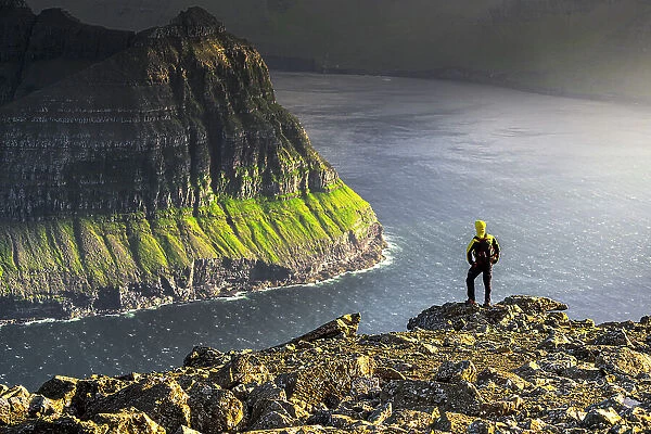 One man admiring the majestic cliffs along a fjord standing on top of mountain during a hike, Vidoy Island, Faroe Islands, Denmark, Europe
