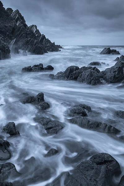 Swirling waves over dark rocks in a rugged North Devon cove, Devon, England, United