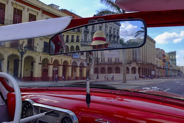 Taxi driver in straw hat seen in rear-view mirror of vintage car, Havana, Cuba