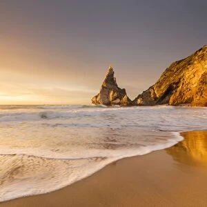 Golden reflections of the cliffs on Praia da Ursa beach bathed by ocean at sunset