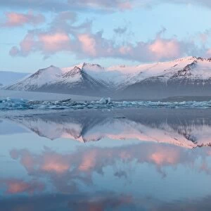 Panoramic view across the calm water of Jokulsarlon glacial lagoon towards snow-capped mountains and icebergs bathed in the last light of a winters afternoon, at the head of the Breidamerkurjokull Glacier on the edge of the Vatnajokull National Park, South Iceland, Iceland, Polar Regions