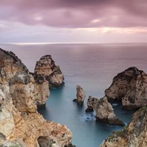Photographer on top of cliffs surrounded by sea under the pink sky at sunrise, Ponta Da Piedade