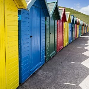 Row of colourful beach huts and their shadows with green hill backdrop, West Cliff Beach
