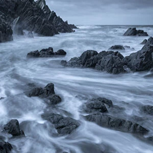 Swirling waves over dark rocks in a rugged North Devon cove, Devon, England, United