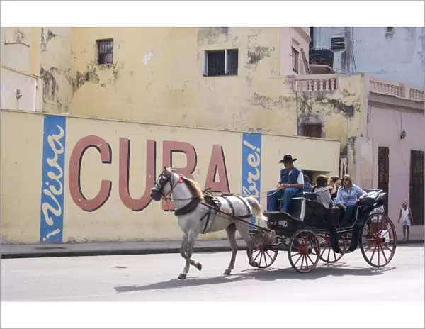 Tourists in a horse carriage passing a wall sign saying Live free Cuba