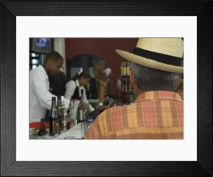 Framed Print : Back view of a man in a straw hat sitting at a bar, with bar staff in the background Framed Print : Back view of a man in a straw hat sitting at a bar, with bar staff in the background
