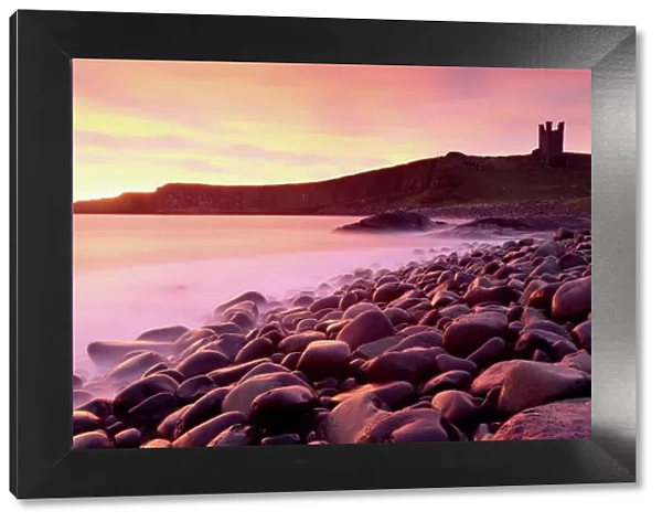 An imposing silhouette of Dunstanburgh Castle against a magnificent sky at sunrise with a beach of basalt boulders in the foreground, Embleton Bay, near Alnwick, Northumberland, England, United