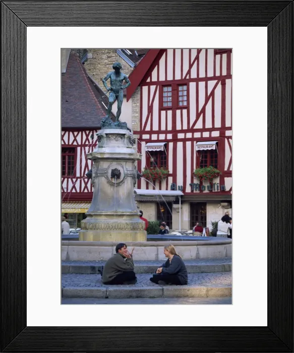 Framed Print : A couple sit in front of a fountain in the Place Francois Rude, with half timbered houses behind Framed Print : A couple sit in front of a fountain in the Place Francois Rude, with half timbered houses behind