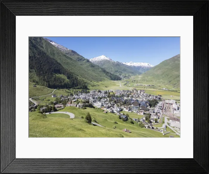 Framed Print : The alpine village of Andermatt surrounded by green meadows, and snowy peaks in the background Framed Print : The alpine village of Andermatt surrounded by green meadows, and snowy peaks in the background