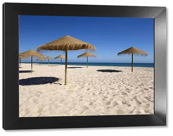 Straw umbrellas on empty white sand beach with clear sea behind, Ilha do Farol, Culatra