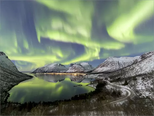 Northern lights (aurora borealis) and stars light up the snowy peaks reflected in the cold sea