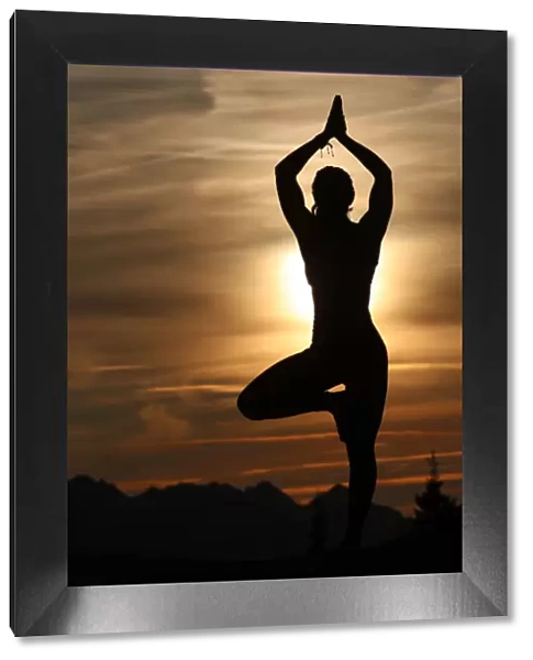 Silhouette of a woman in Vrkasana (tree pose) practising yoga against the light of the