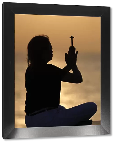 Silhouette of faithful woman praying with Christian cross at sunset as concept for