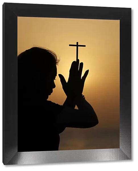 Silhouette of faithful religious woman praying with Christian cross at sunset