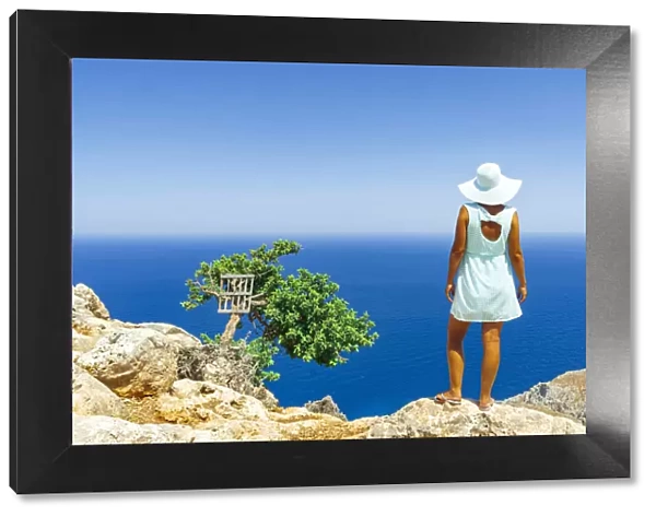 Back view of woman with fashion dress and hat looking at the sea from cliffs, Crete