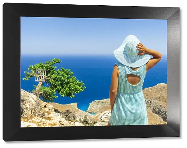 Back view of young woman with dress and sun hat watching the sea, Crete, Greek Islands