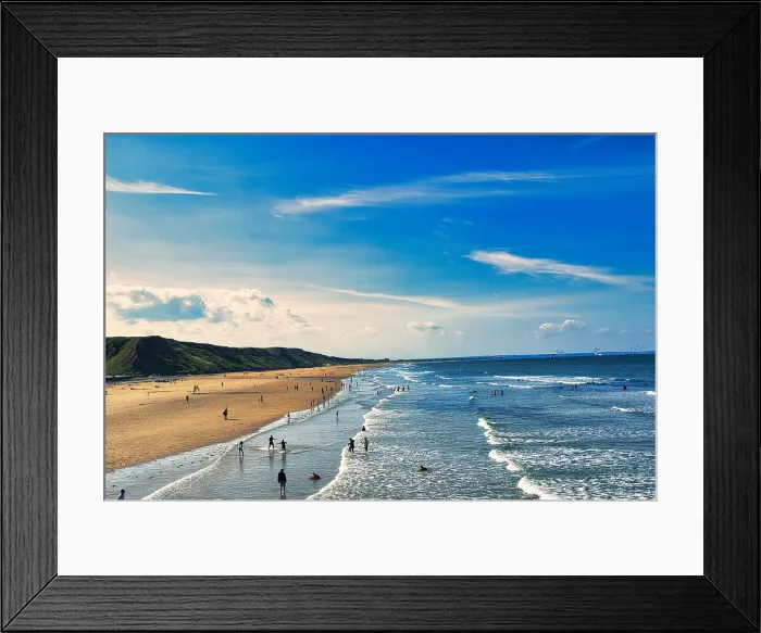Framed Print : Sunny beach scene with people and rolling waves, blue sky with clouds overhead, and a hilly backdrop in Saltburn-by-the-Sea, England Framed Print : Sunny beach scene with people and rolling waves, blue sky with clouds overhead, and a hilly backdrop in Saltburn-by-the-Sea, England