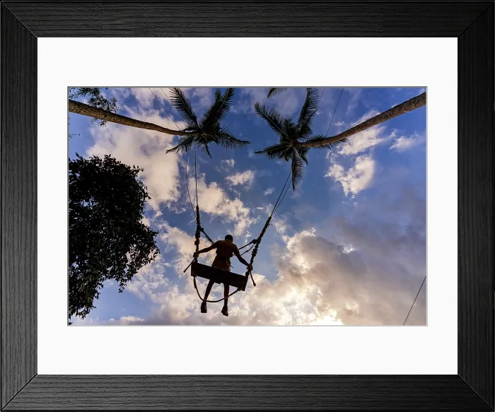 Framed Print : Low angle view of a cheerful boy having fun on a jungle swing between palm trees against clouds at sunset, Ubud, Bali, Indonesia