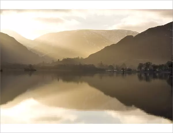Morning sunlight bursting through clouds over fells with reflections in Lake Ullswater