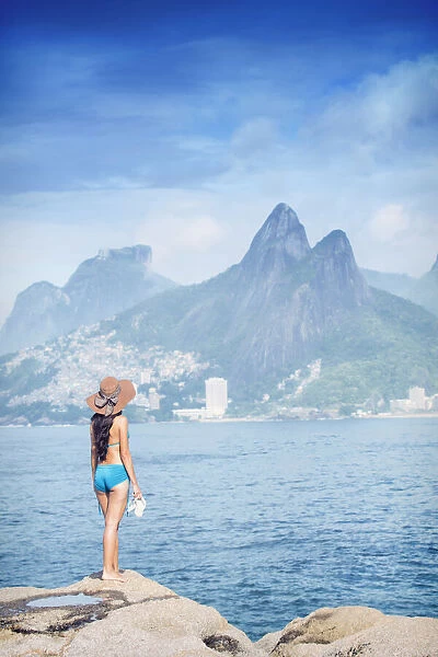 A 20-25 year old young Brazilian woman standing on the Arpoador rocks with Ipanema