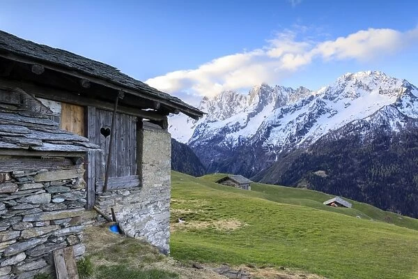 Alpine hut framed by meadows and snowy peaks at dawn, Tombal, Soglio, Bregaglia Valley
