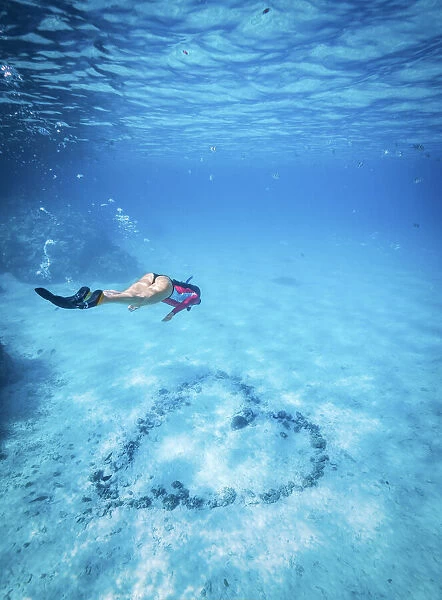 An athletic woman free-dives to inspect a heart arranged with rocks on the bottom of