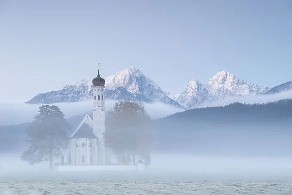 The autumn fog at sunrise frames St. Coloman Church surrounded by snowy peaks, Schwangau