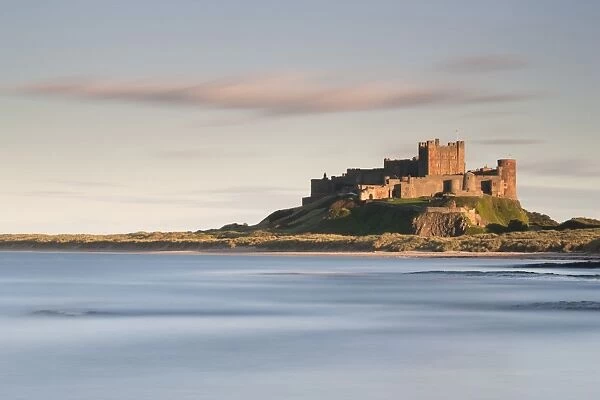 Bamburgh Castle bathed in golden evening light overlooking Bamburgh Bay with the sea filling the foreground, Northumberland, England, United