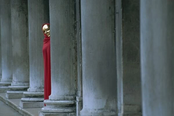 Carnival model in red cape and gold mask peering from columns in St