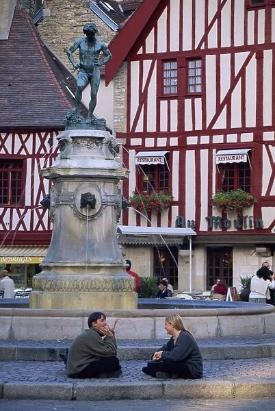 A couple sit in front of a fountain in the Place Francois Rude, with half timbered houses behind