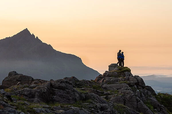 A couple standing on a rocky peak watching a mountain sunset with Sgurr nan Gillean in