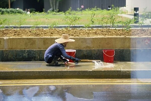 An elderly woman in a straw hat washes the pavement