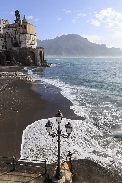 Fancy street lamp, rusty anchor and wave breaking on beach, distant church, Atrani