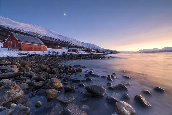 Fiery sky at sunset and typical Rorbu, snowy peaks and frozen sea, Djupvik, Lyngen Alps