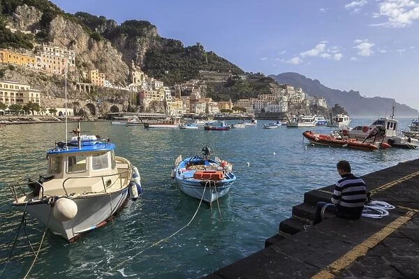 Fisherman working on harbour quayside with view towards Amalfi town and fishing boats