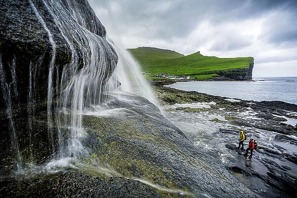 High angle view of two hikers admiring a majestic waterfall standing on cliffs, Gjogv, Eysturoy Island, Faroe Islands, Denmark, Europe