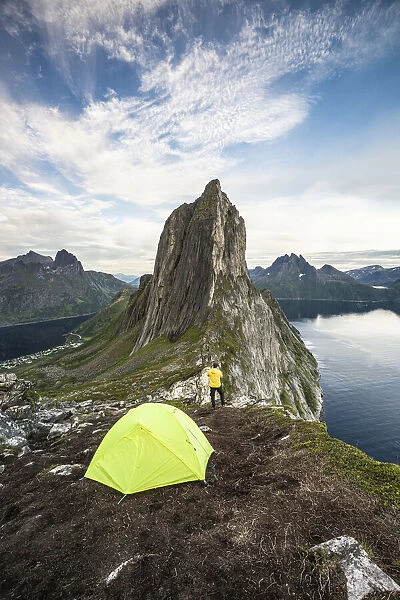 Hiker man with camping tent admiring Mount Segla and fjord from Hesten peak at sunset