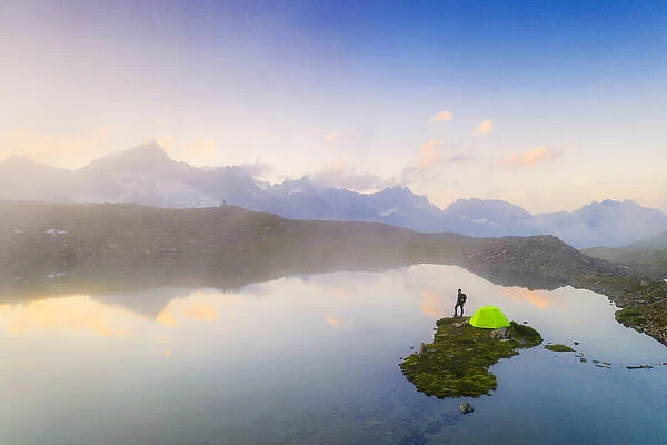 Hiker man with tent admiring a misty sunrise over mountains from Obere Schwarziseeli lake