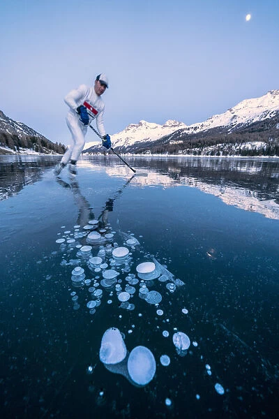 Ice hockey player man skating on Lake Sils covered in ice bubbles at dusk, Engadine