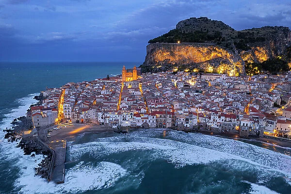 The illuminated fishing village of Cefalu with the rock on top of the old town seen from the drone at dusk, Cefalu, Palermo province, Tyrrhenian Sea, Sicily, Italy, Mediterranean, Europe