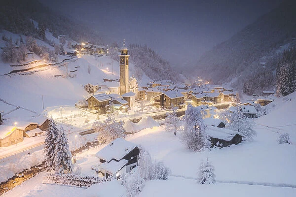 Illuminated village of Gerola Alta and frozen river covered with snow during winter dusk