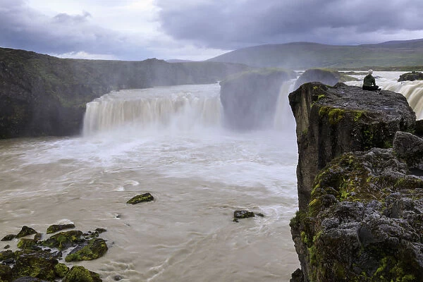 Lady sits cross legged on a rock overlooking Godafoss (Waterfall of the Gods), long exposure