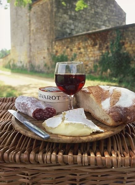 Still life of bread, glass of red wine, cheese and sausage, picnic meal on top of a wicker basket