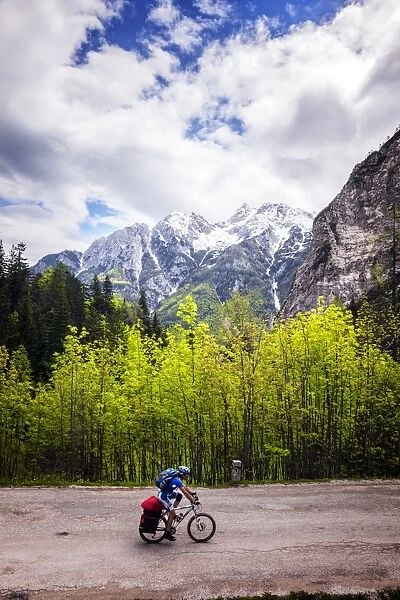 A lone cyclist travels along a mountain road with trees and the Julian Alps in the background
