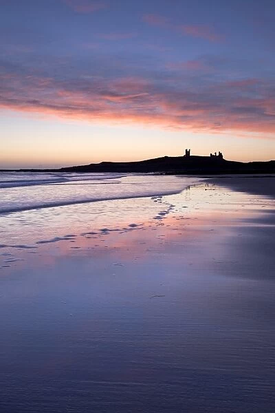 Looking across Embleton Bay at sunrise towards the silhouetted ruins of Dunstanburgh Castle in the distance and the vivid colours in the sky reflecting in the sea and wet sand, Embleton, near Alnwick, Northumberland, England, United Kingdom